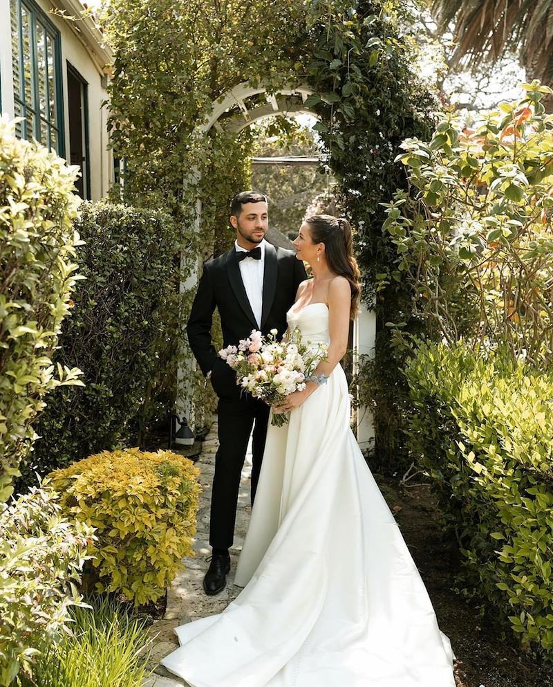A happy bride and groom pose together in front of a charming house, radiating love and joy on their special day.