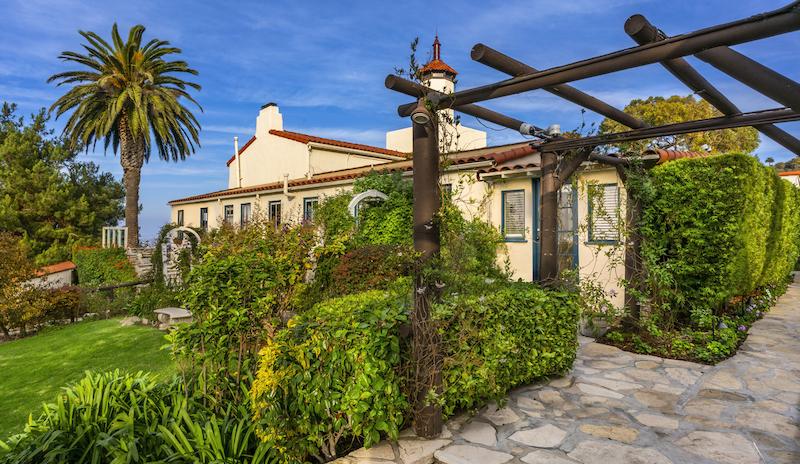 A Spanish-style villa with lush gardens, a palm tree, and a clear blue sky.