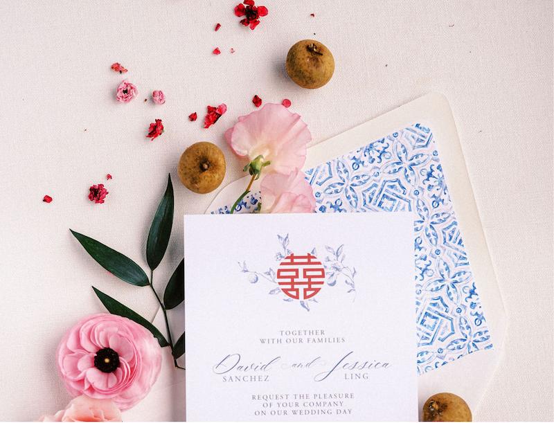 Wedding invitation card with floral design, surrounded by pink flowers, green leaves, three small round fruits, and red petals on a light background. An envelope with a blue geometric pattern is underneath.
